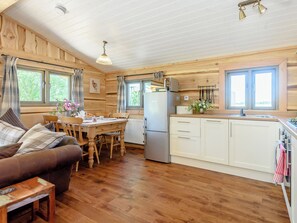 A cozy cabin interior with a wooden dining table, a sofa, a kitchen area with a refrigerator, and a window with curtains. - 2 bedroom accommodation in Croft, near Skegness (Croft, near Skegness)