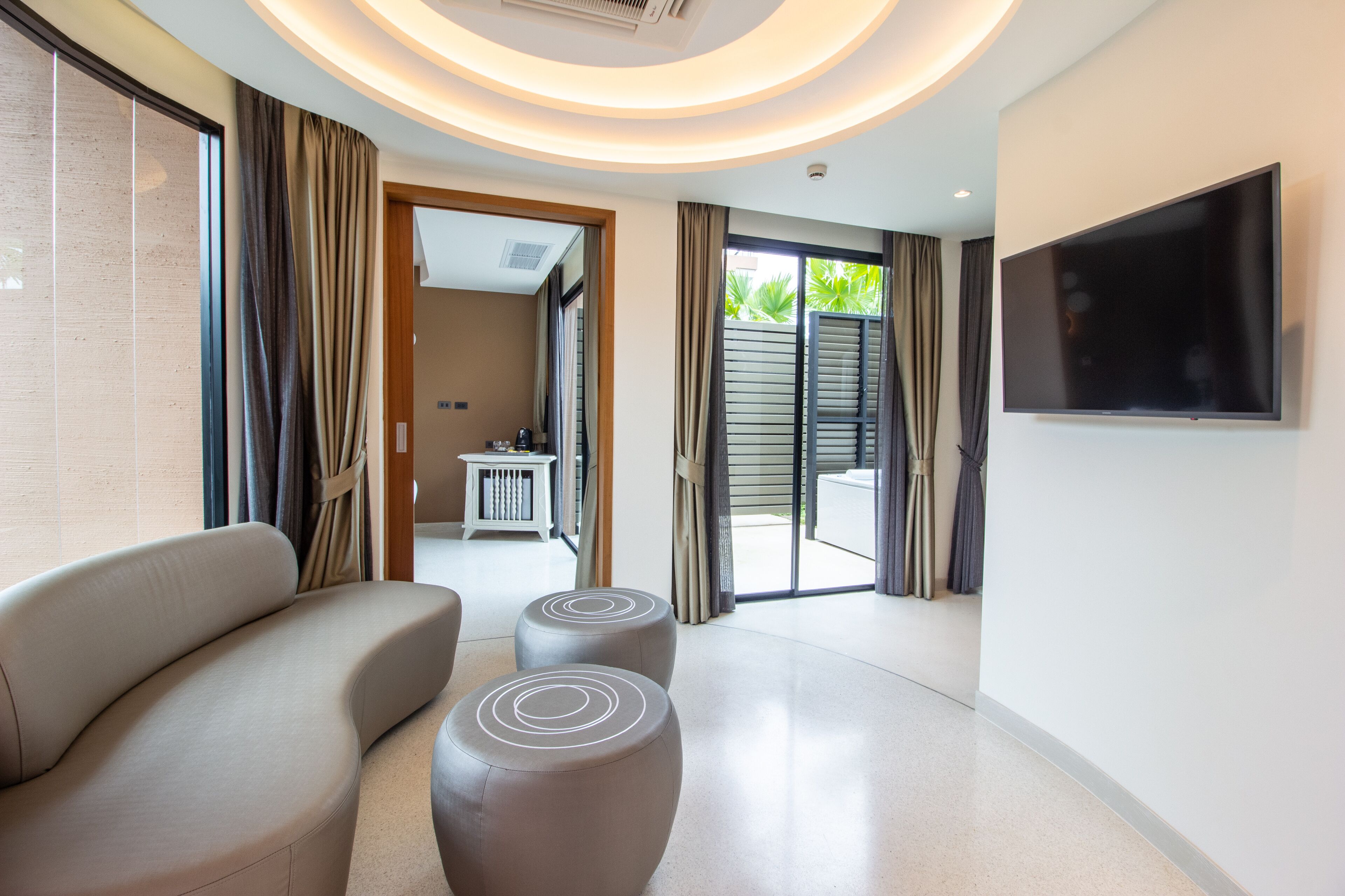 A modern hotel room with a sofa, ottomans, a flat-screen TV, and a view of the outdoor area.