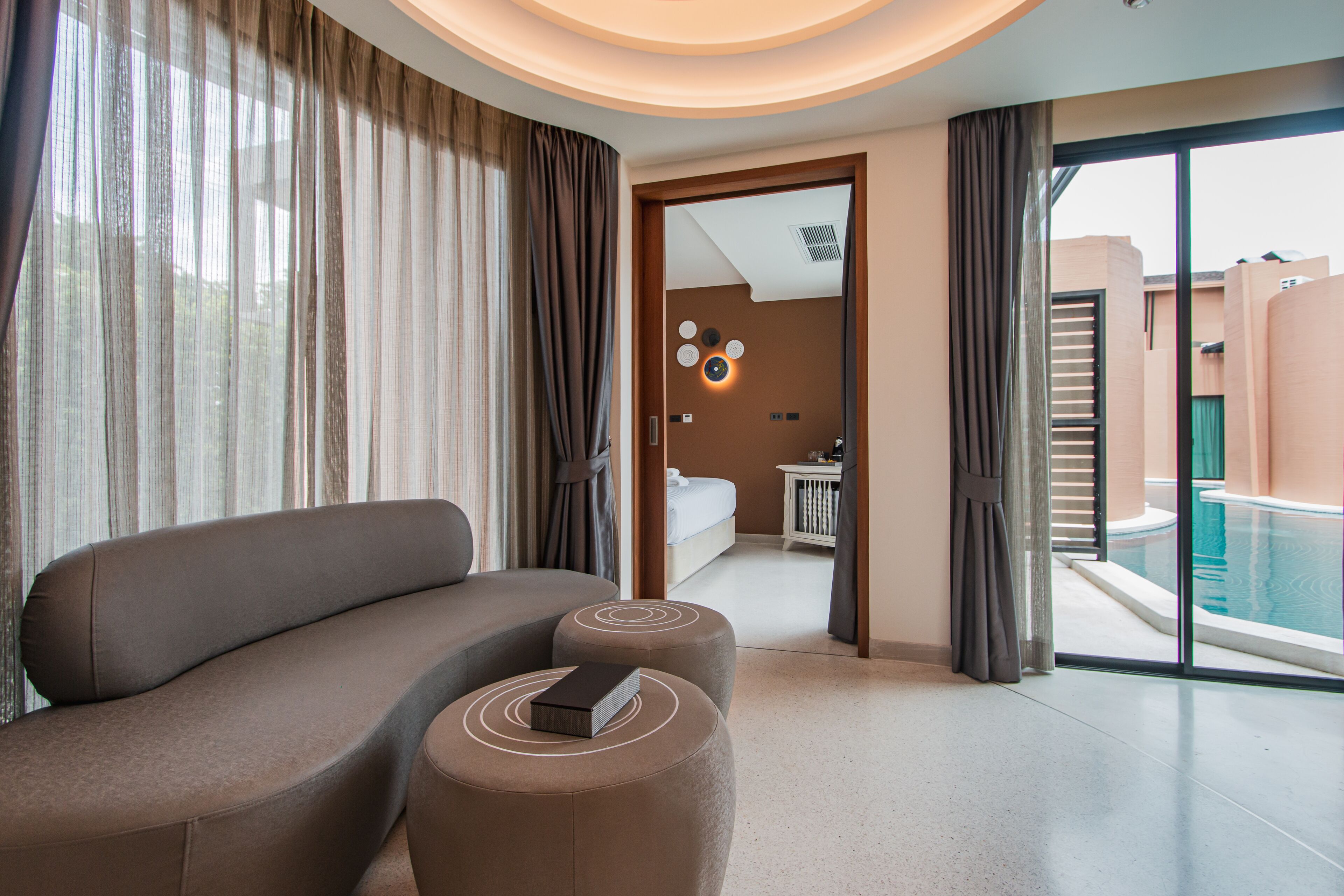 A modern hotel room with a curved sofa, ottoman, and a view of a swimming pool.