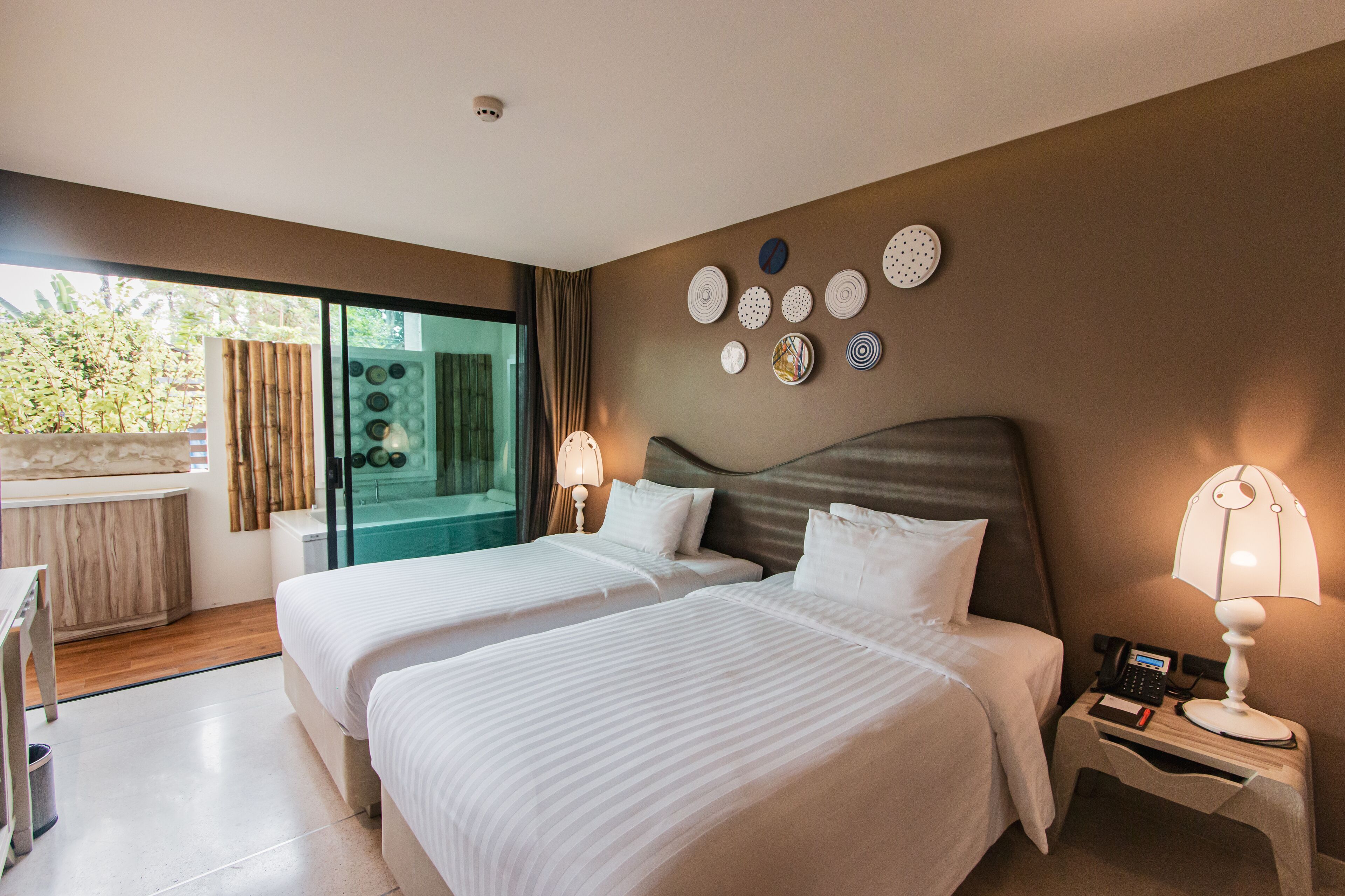 A hotel room with two beds, a bathroom with a glass shower, and a balcony with a view of trees.