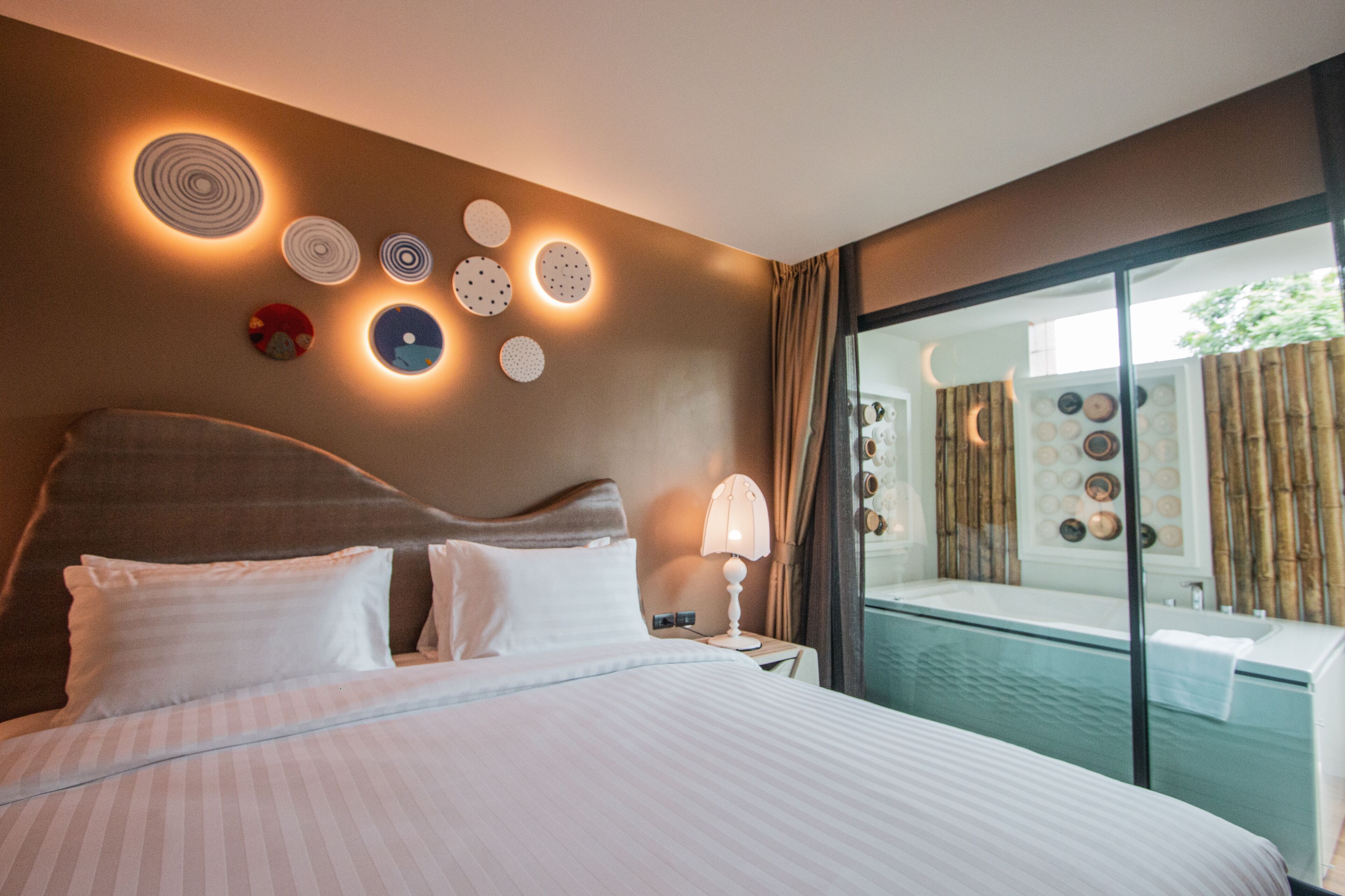 A hotel room with a large bed, a bedside table with a lamp, and a view of a pool area through a glass door.