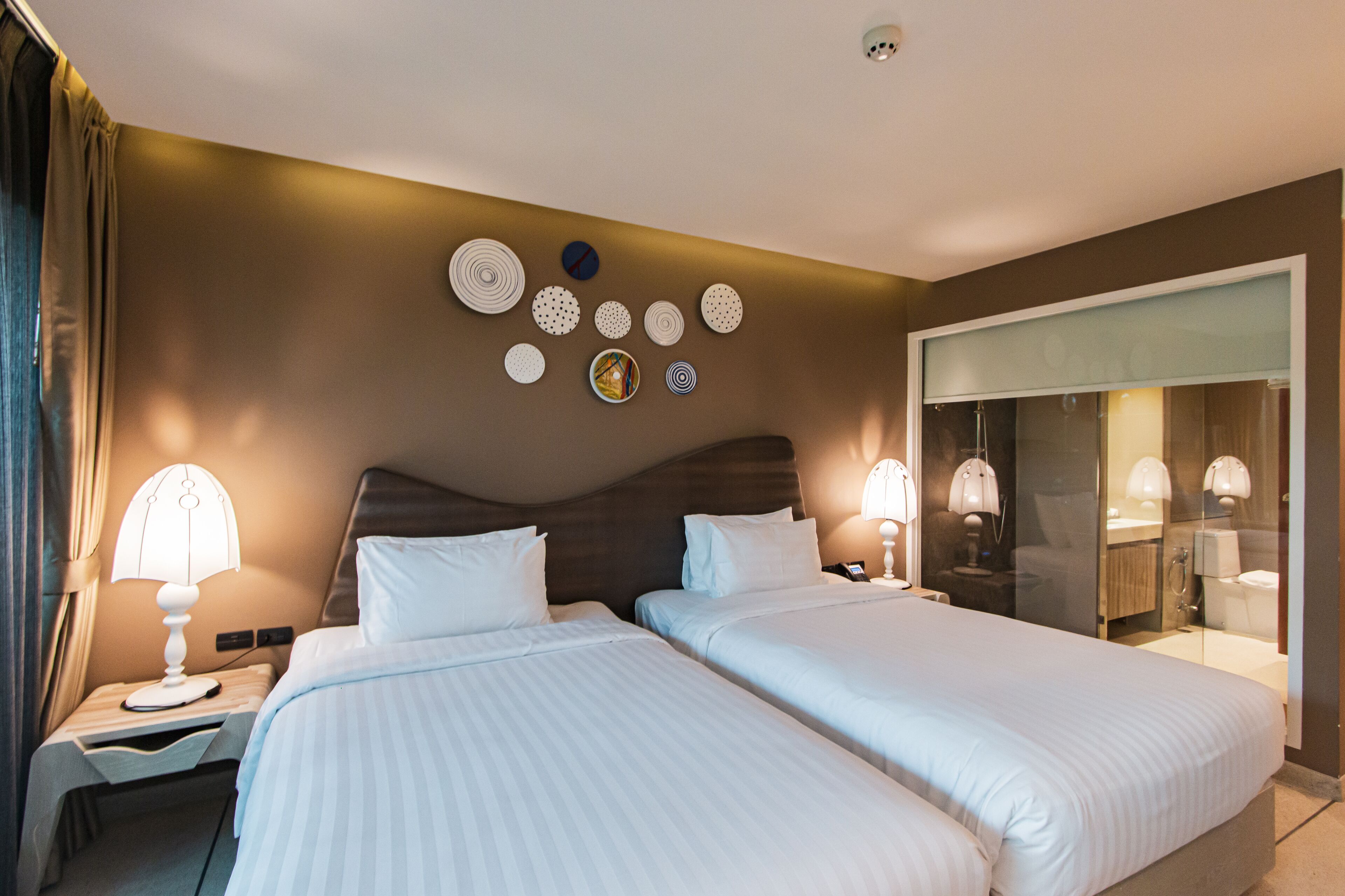 A hotel room with two beds, a nightstand with a lamp, a wall with decorative plates, and a bathroom visible through a glass door.