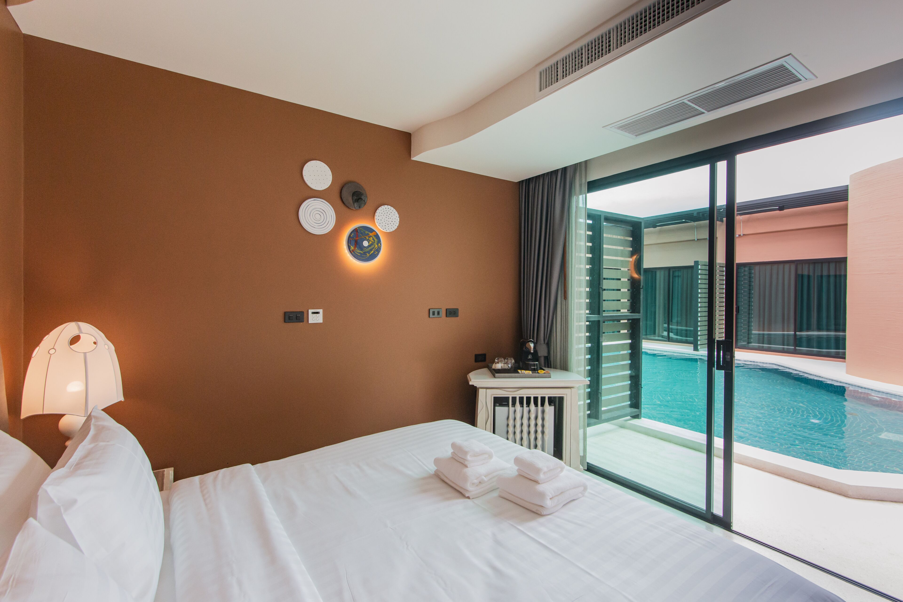 A hotel room with a bed, a nightstand, a lamp, a wall with decorative plates, and a view of an indoor pool through a glass door.