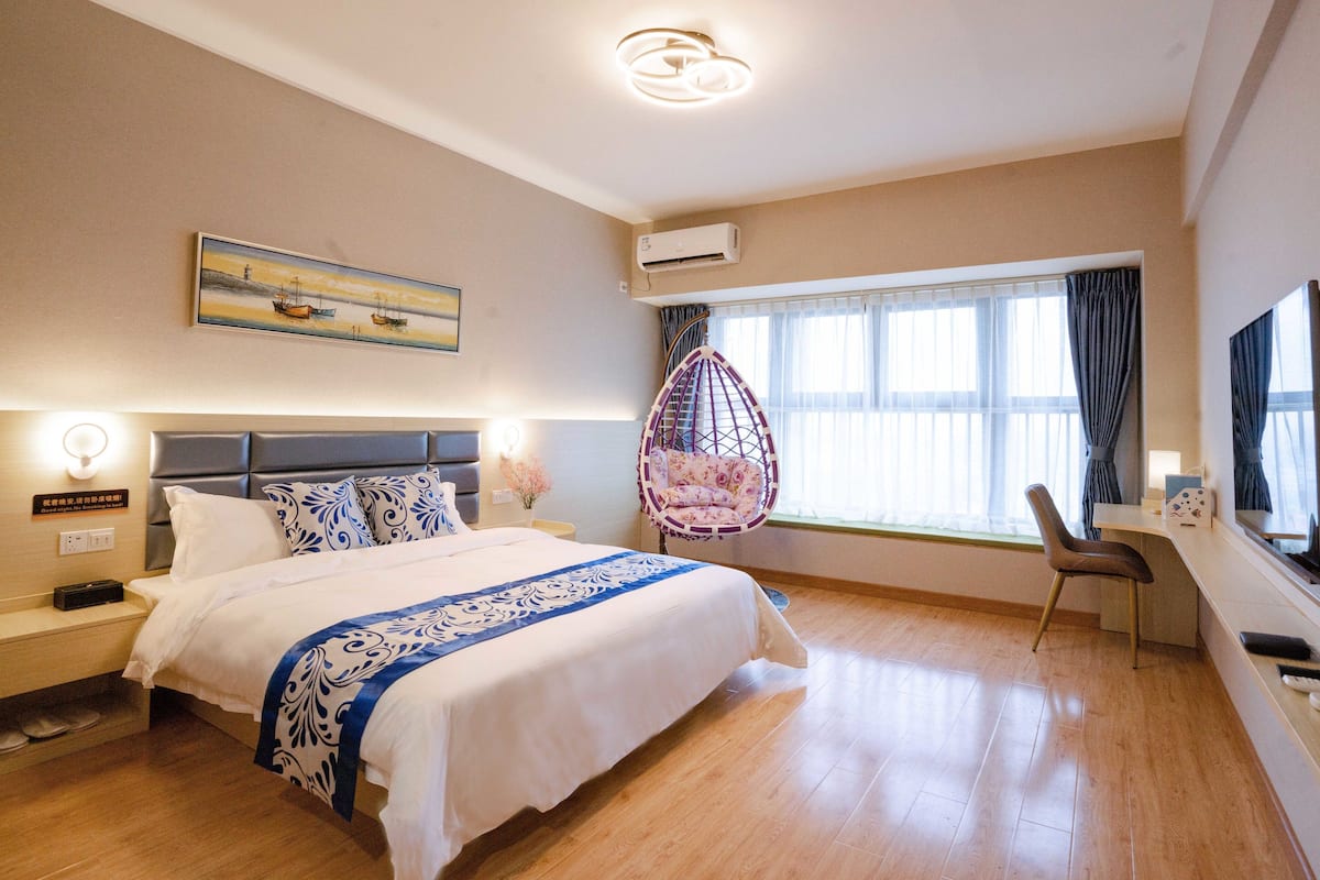 A hotel room with a bed, a hanging chair, a desk with a TV, and a large window with curtains.