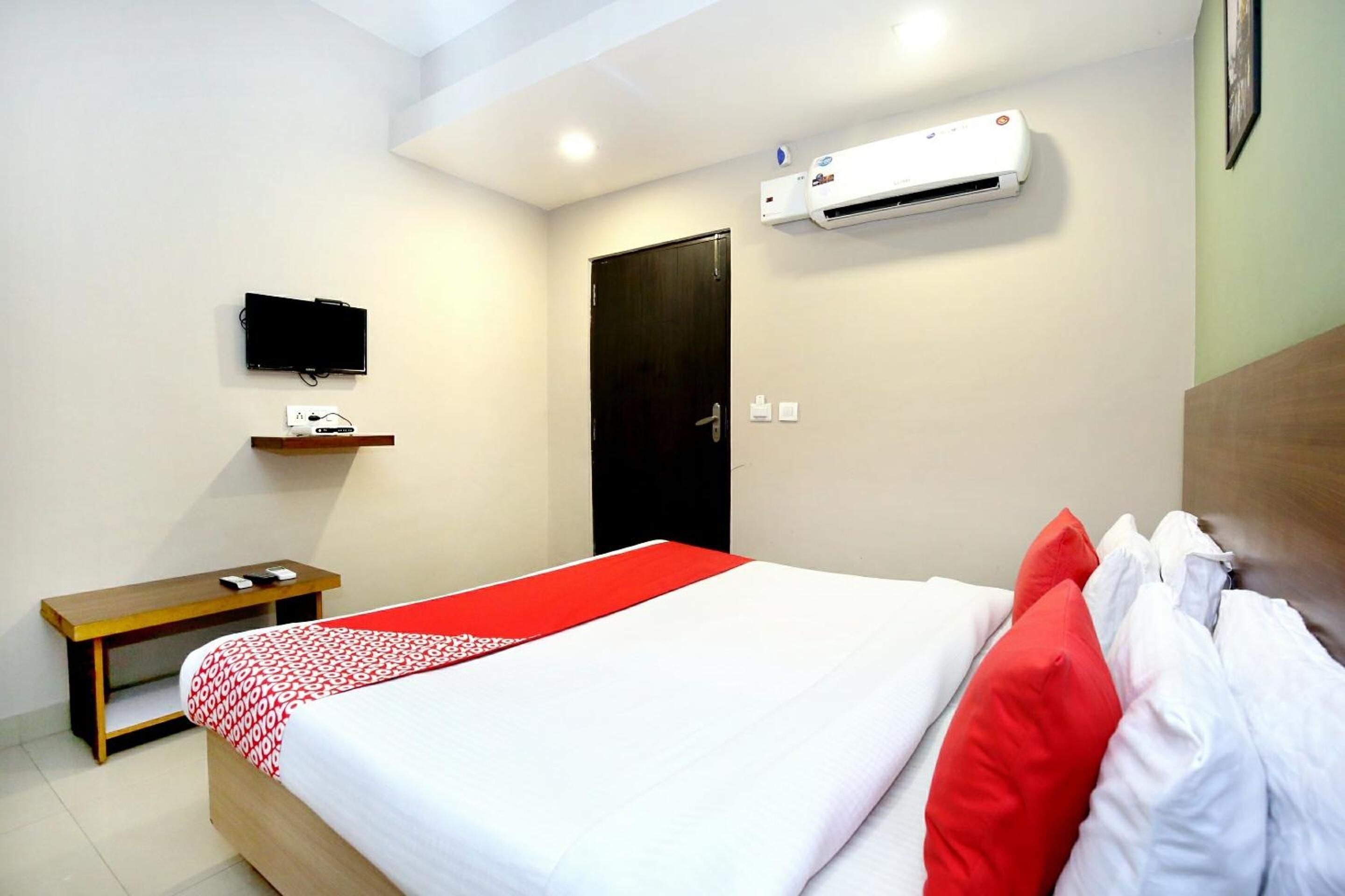 A hotel room with a bed, a bedside table, a TV mounted on the wall, and an air conditioning unit.