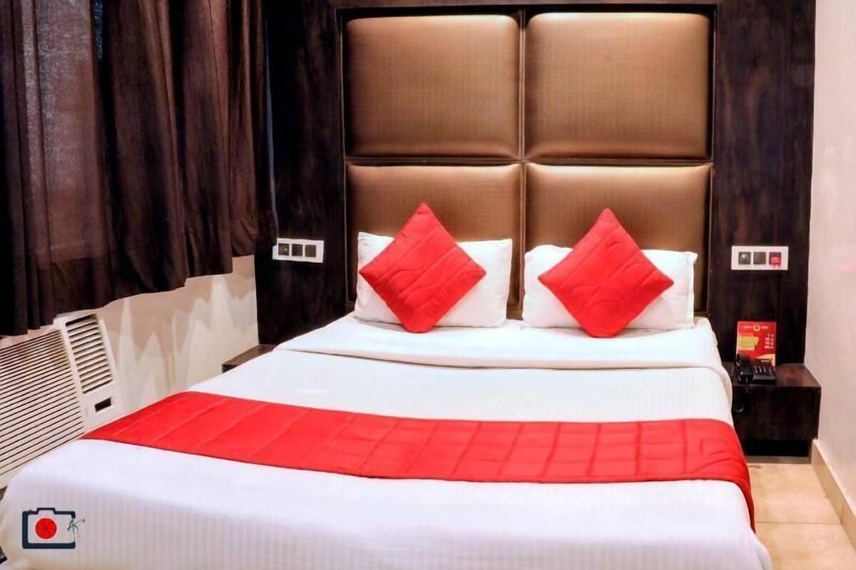 A hotel room with a large bed, dark curtains, a headboard with a padded design, and a red accent pillow.