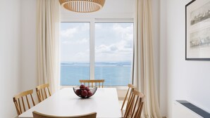 Un comedor con una mesa blanca y sillas de madera, un bol de fruta, y vista al mar a través de amplios ventanales.