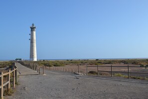 A lighthouse with a wooden walkway leading to it, surrounded by a sandy landscape and a clear sky. - Atlántico Home page Morro Jable (Morro Jable)