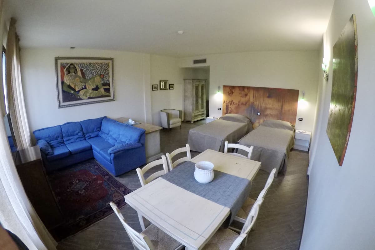 Une chambre d’hôtel avec un canapé bleu, une table à manger avec des chaises, deux lits avec des têtes de lit, un tableau au mur et une petite table avec un vase.