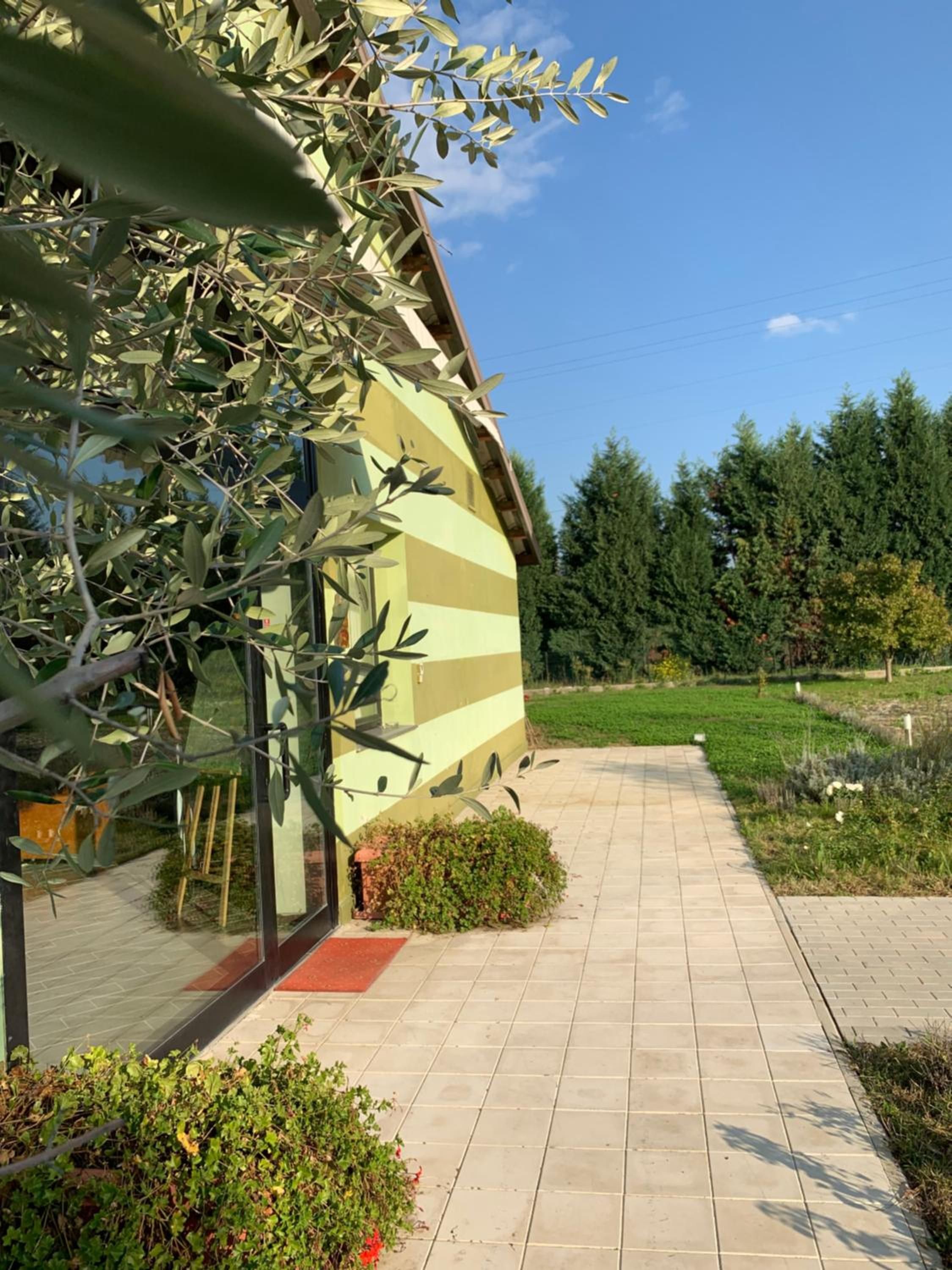 Ein Gebäude mit einer gestreiften Fassade, ein gepflasterter Weg davor und ein Garten mit verschiedenen Pflanzen und Bäumen.