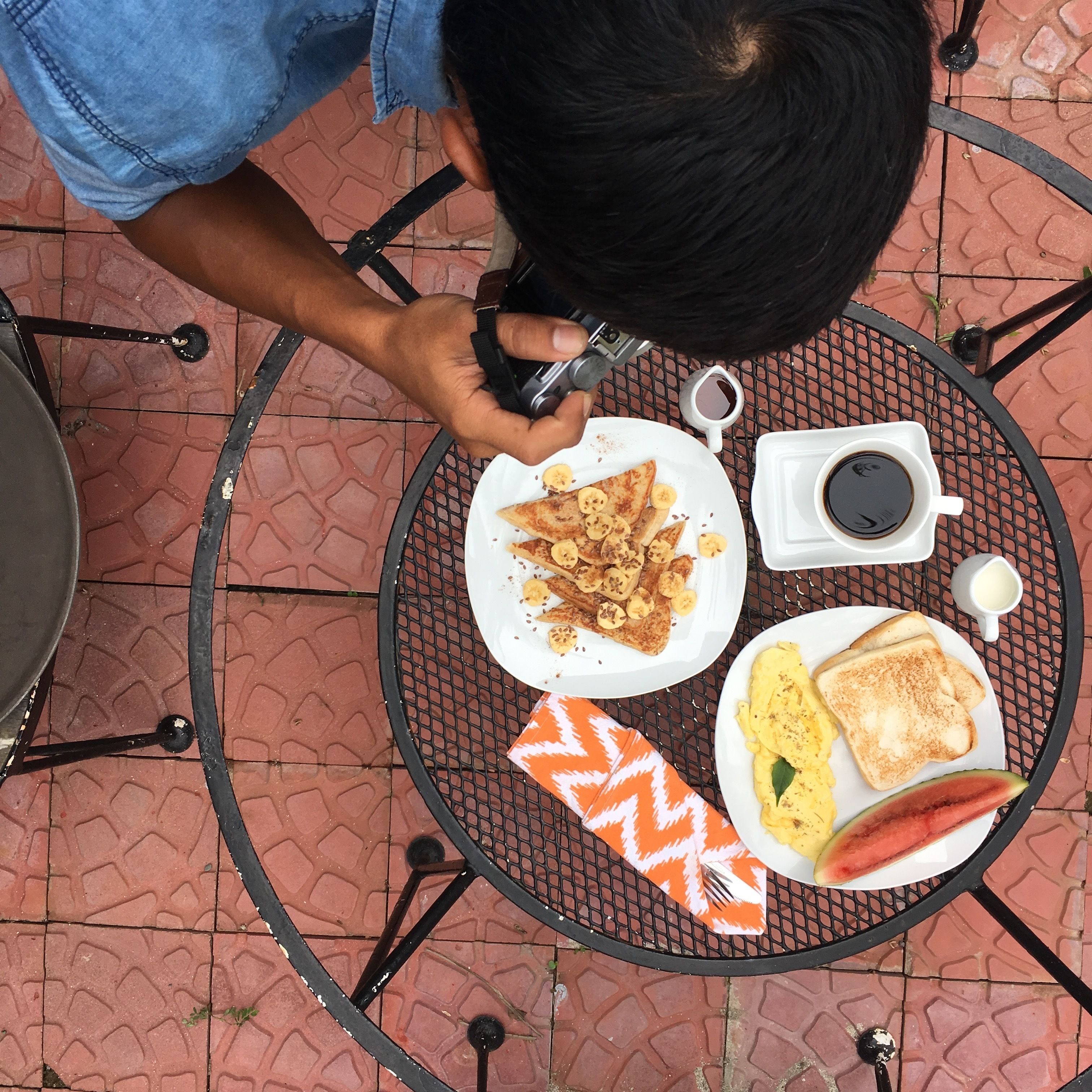A person taking a photo of a meal on a round metal table with a patterned floor.