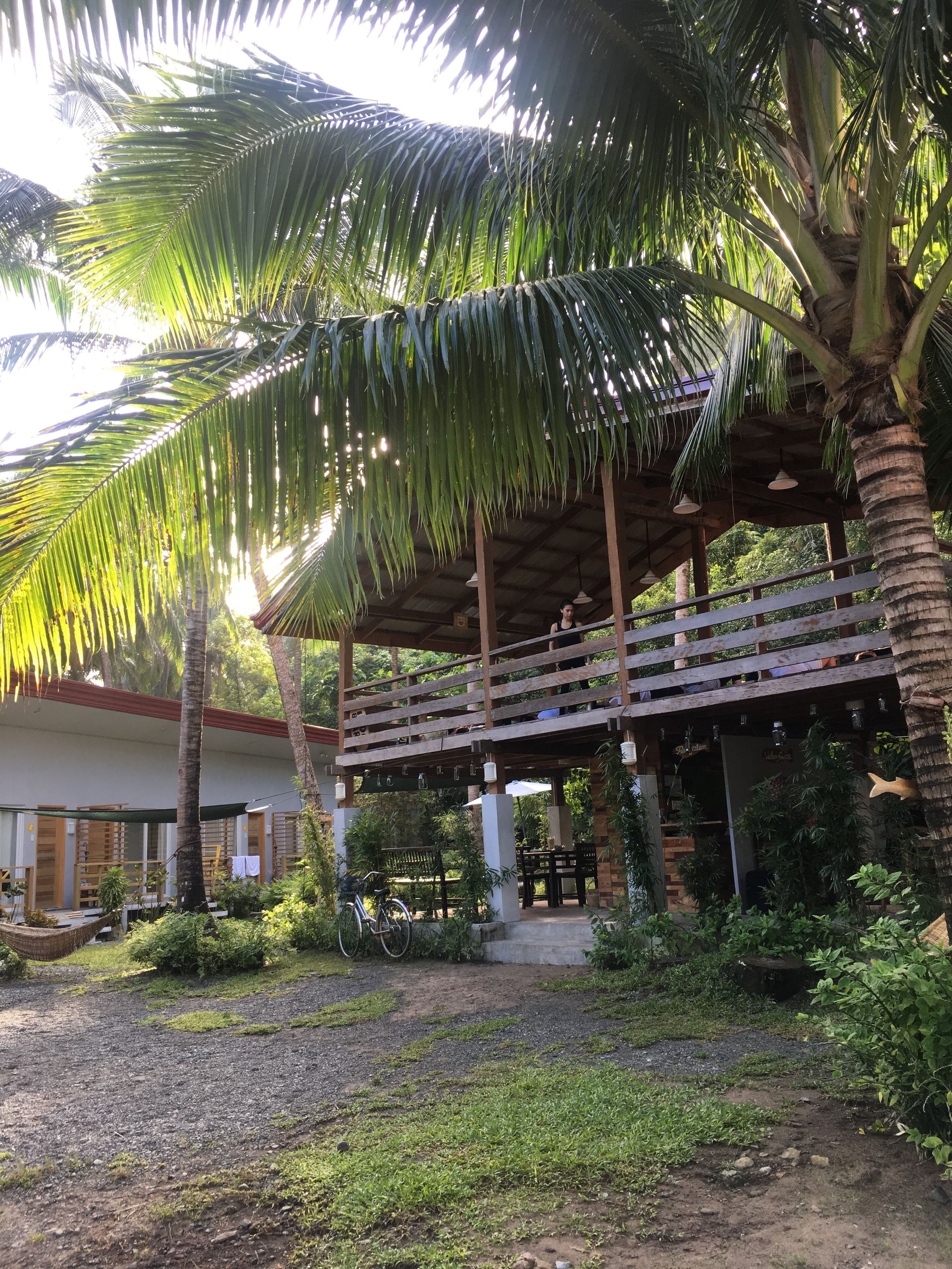 A covered outdoor area with a wooden deck, palm trees, and a building with a sign that reads "Hotel".