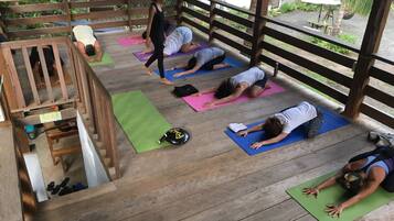 Un groupe de personnes pratiquant le yoga sur des tapis, dans un espace extérieur en bois doté d’une rambarde en bois et agrémenté de verdure en arrière-plan.