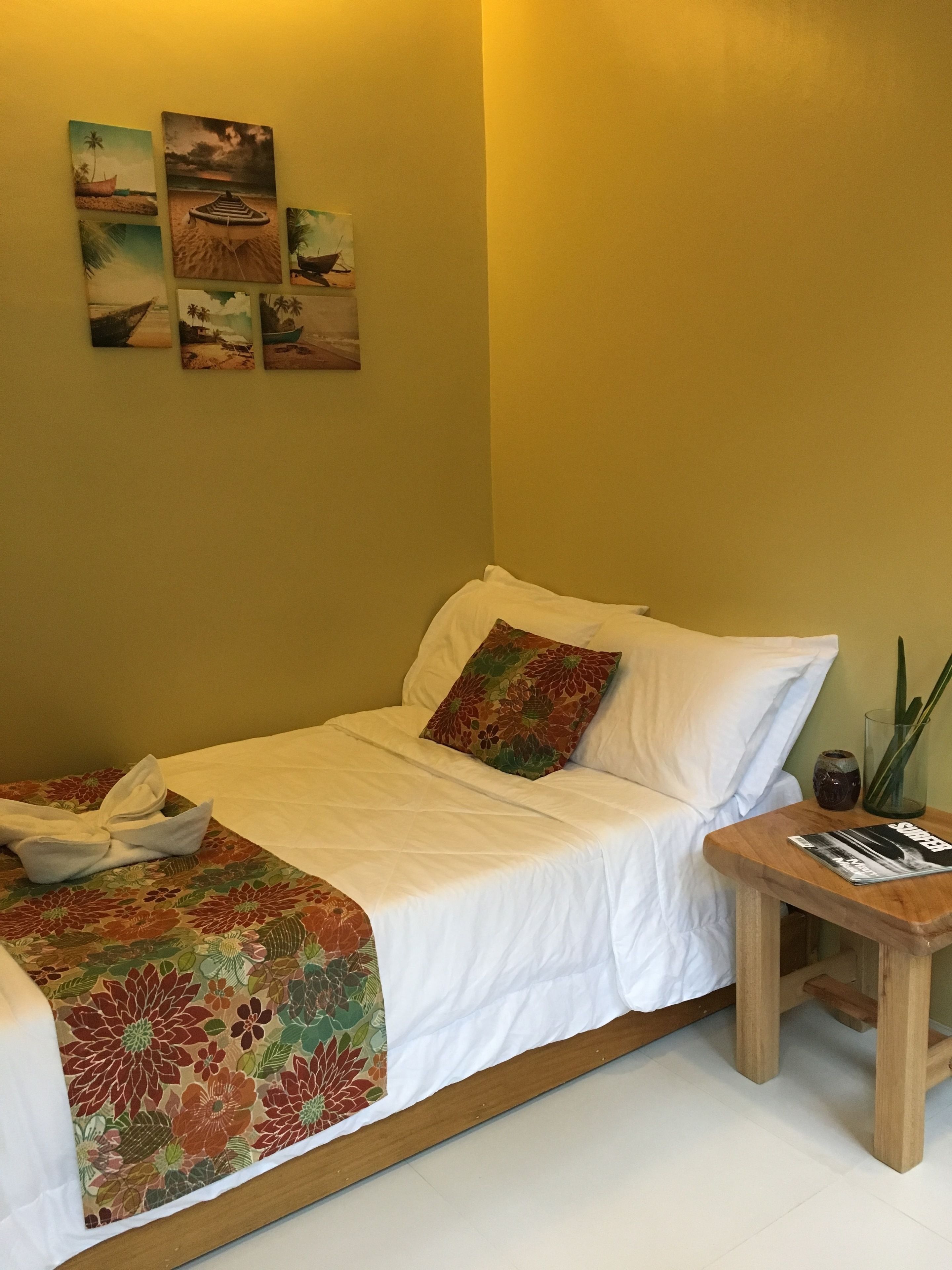 A neatly made bed with a floral bedspread, a wooden nightstand with a magazine, and a wall with multiple paintings.