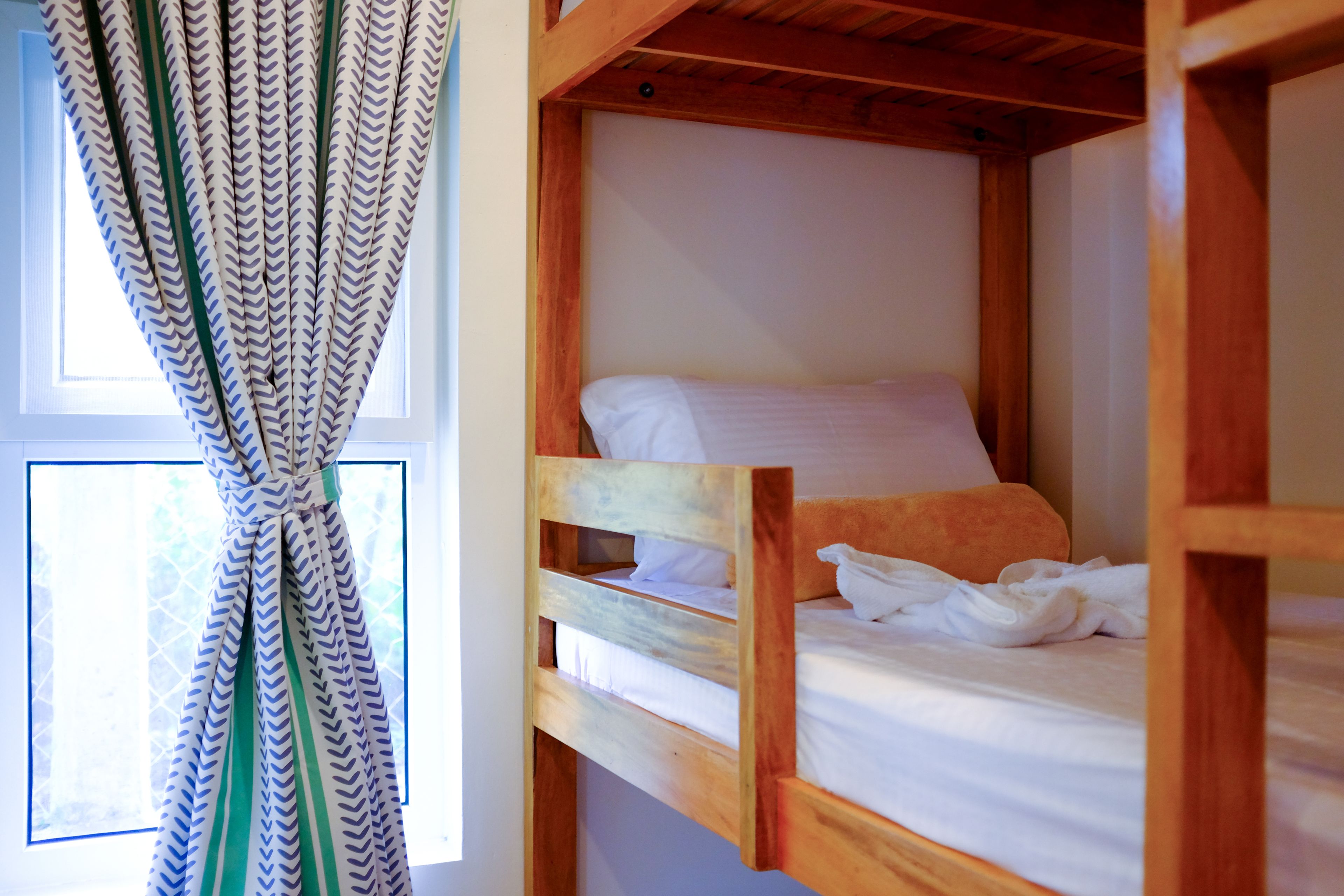 Una cama litera con ropa de cama blanca, una ventana con cortinas y vigas de madera.