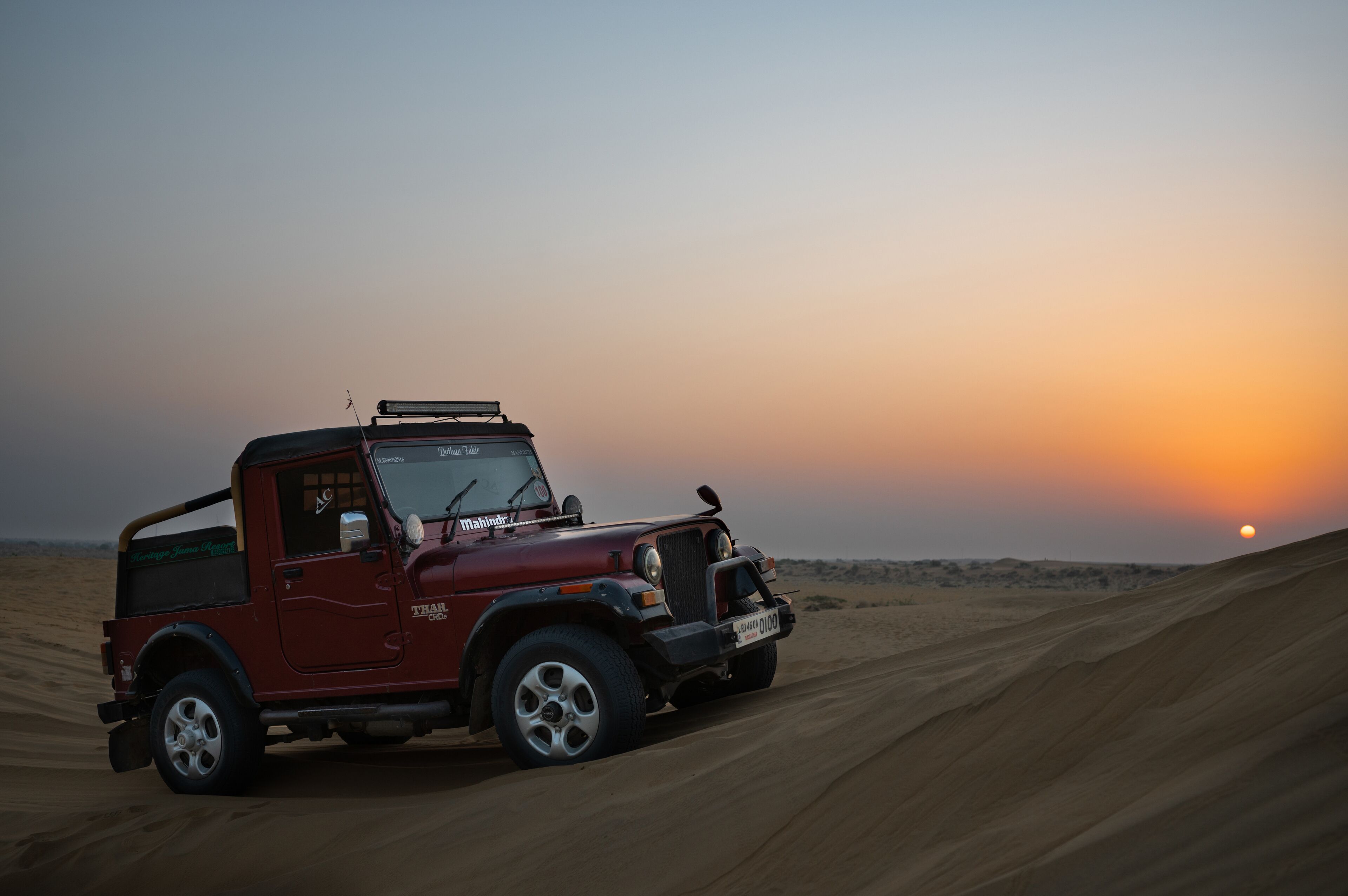 A red Mahindra Thar vehicle on a sandy dune at sunset.
