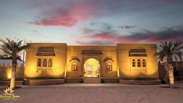 A historic building with arched windows and a central entrance, surrounded by palm trees. - Heritage Juma Resort Camp (Jaisalmer)