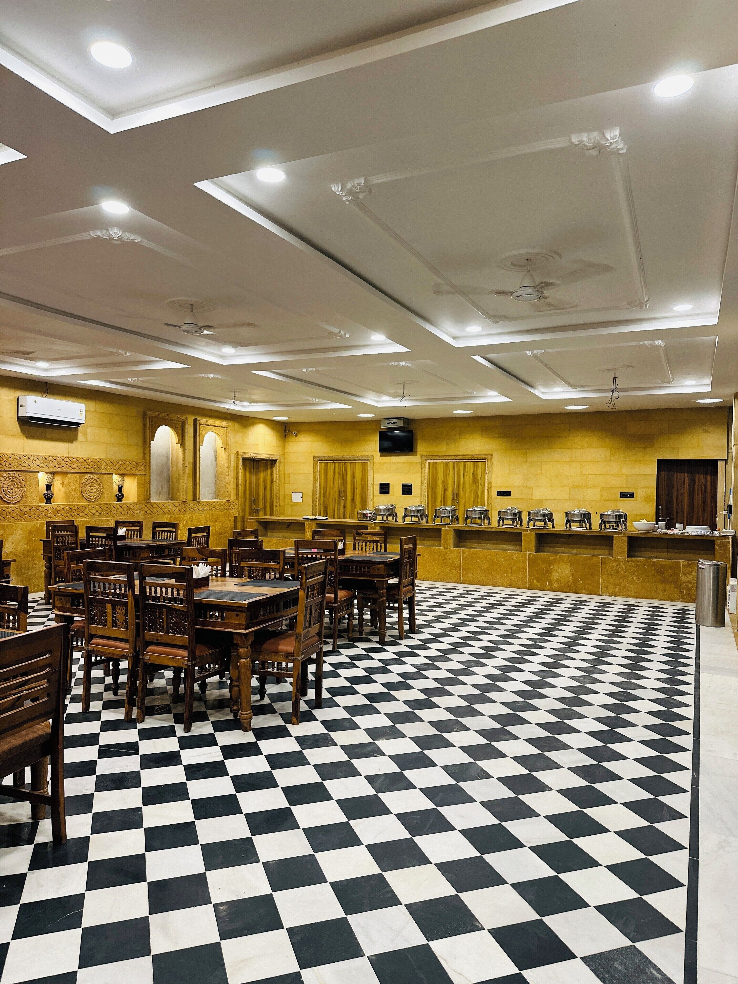A hotel lobby with a checkered floor, wooden tables and chairs, a buffet area, and recessed lighting.