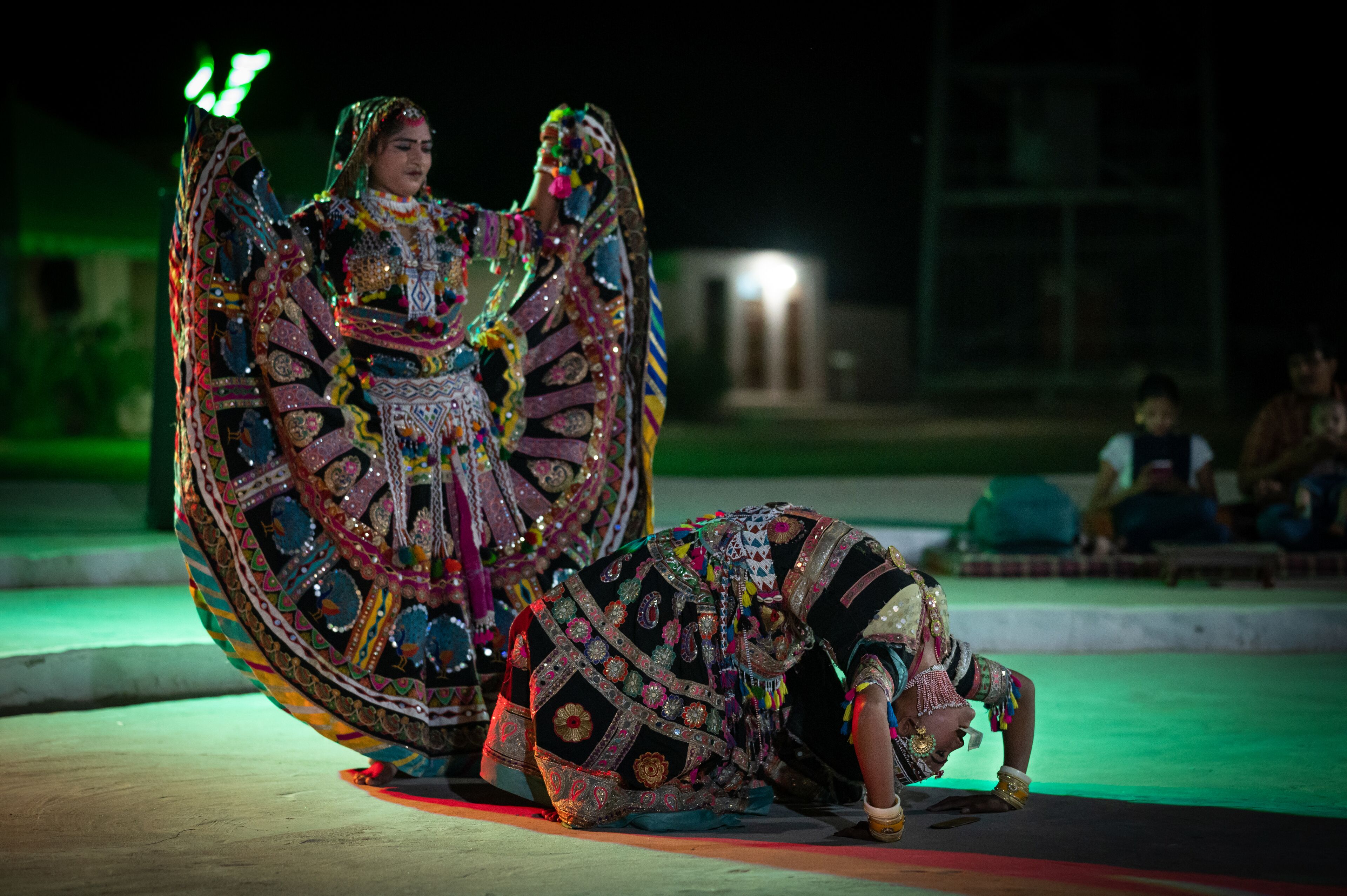 A person in elaborate traditional attire performing on a stage with a backdrop of a building and a crowd in the background.