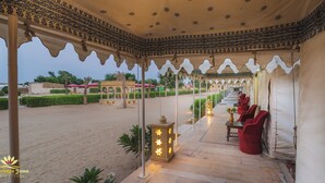 A covered outdoor seating area with patterned ceiling and walls, a sandy ground, and a view of a building in the background. - Heritage Juma Resort Camp (Jaisalmer)