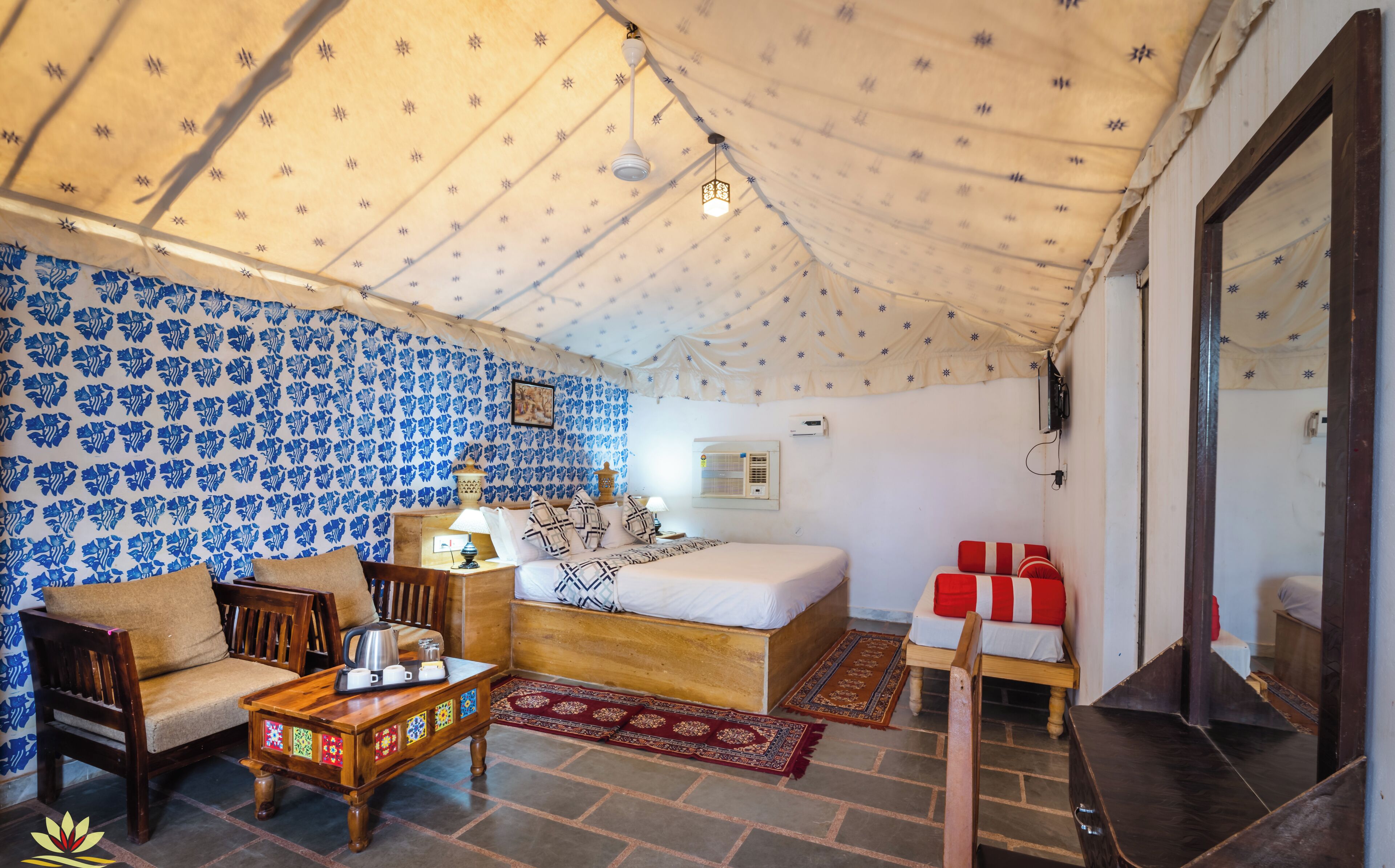 A tent-style room with a bed, a wooden table with cups and a kettle, a chair, and a patterned wall.