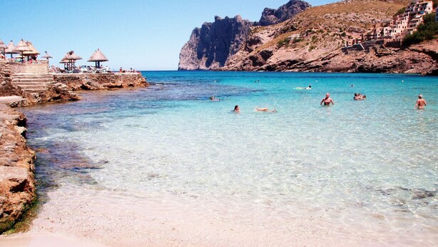 Ein Strand mit klarem türkisblauem Wasser, felsigen Klippen und einigen Sonnenschirmen.