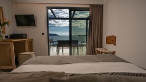 Une chambre d’hôtel avec un grand lit, un bureau équipé d’un ordinateur, une chaise et une vue sur la mer, visible à travers une porte vitrée.