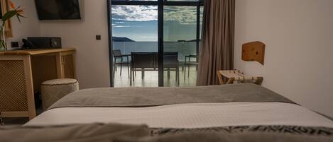 A hotel room with a large bed, a desk with a computer, a chair, and a view of the sea through a glass door.