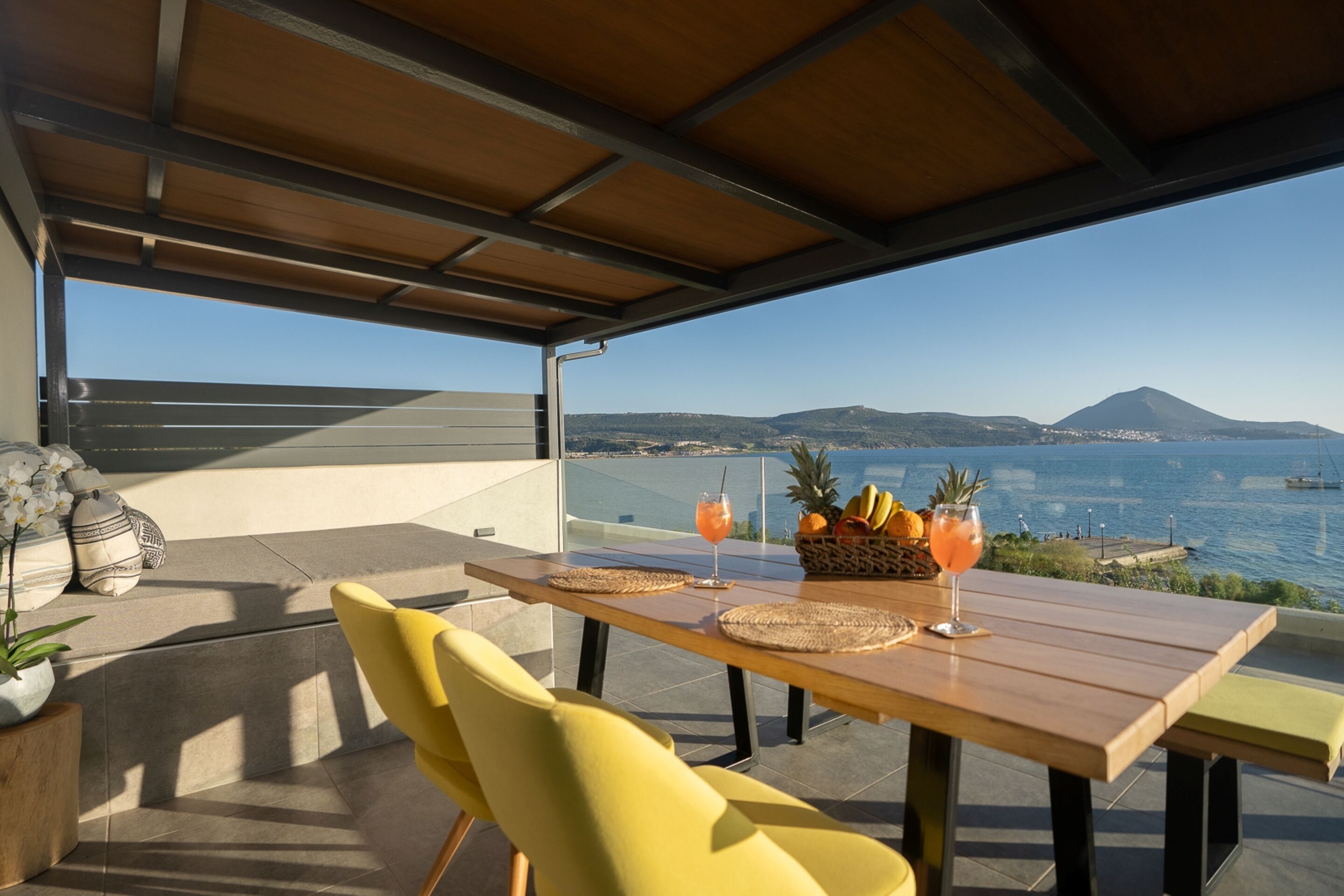 Una mesa de madera con individuales tejidos, sillas amarillas y una cesta de frutas con bebidas en una terraza con vistas a la costa y montañas al fondo.