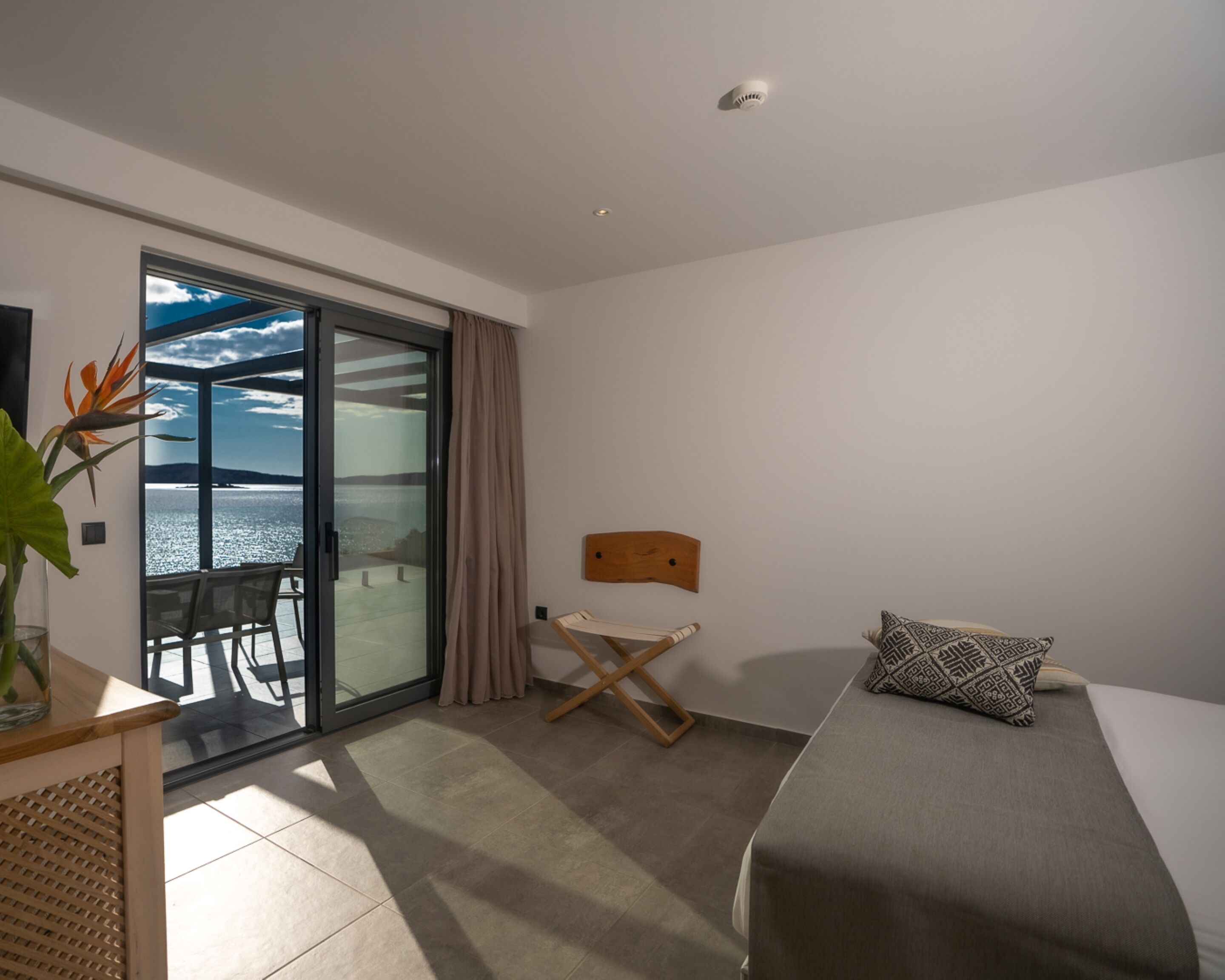 Une chambre d’hôtel moderne équipée d’un lit, d’un petit bureau, d’une chaise, d’une vue sur la mer par une porte vitrée et d’un luminaire au plafond.