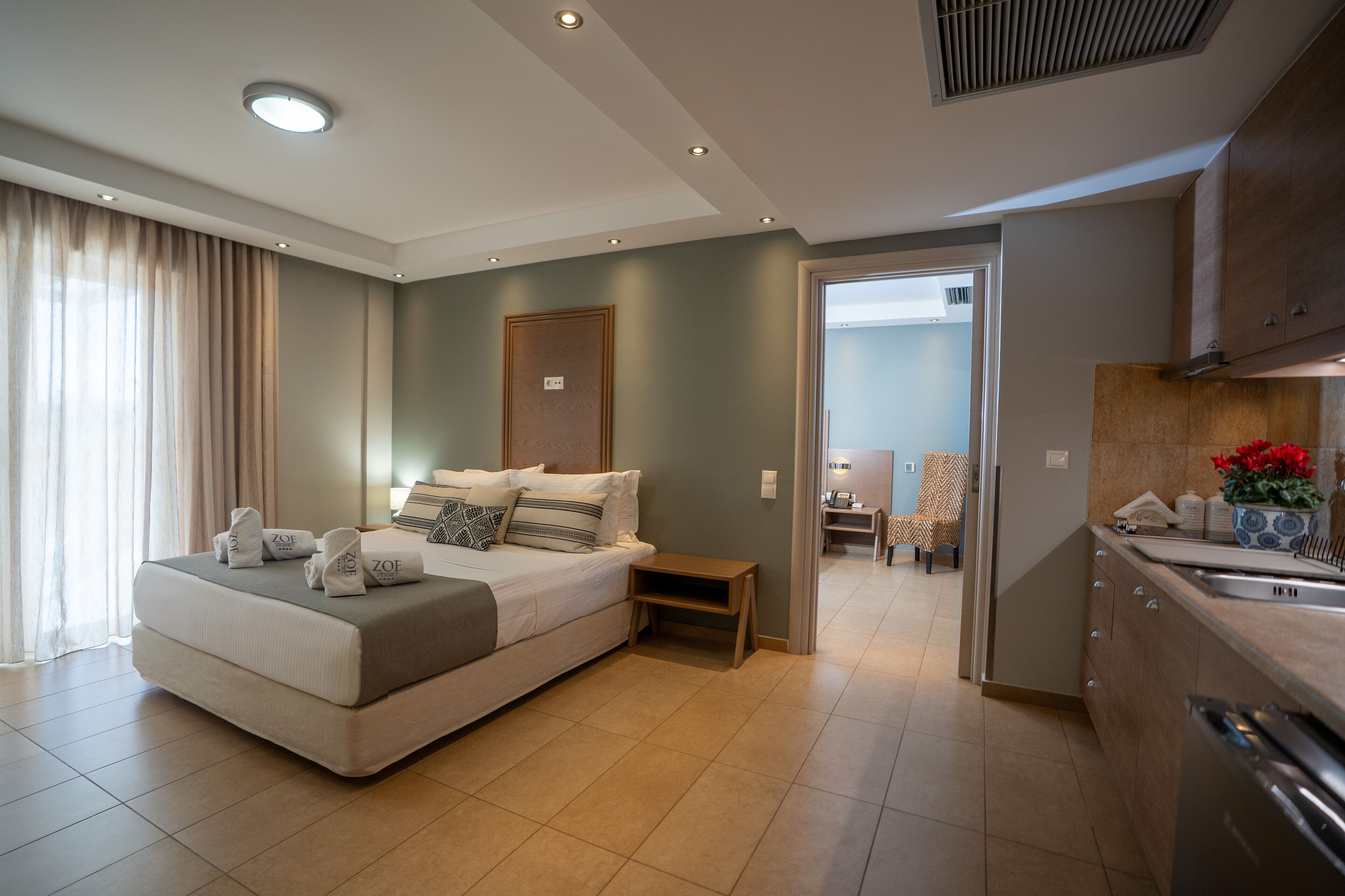 A modern hotel room with a large bed, a small bedside table, a built-in kitchen area, and a view of the adjacent room.