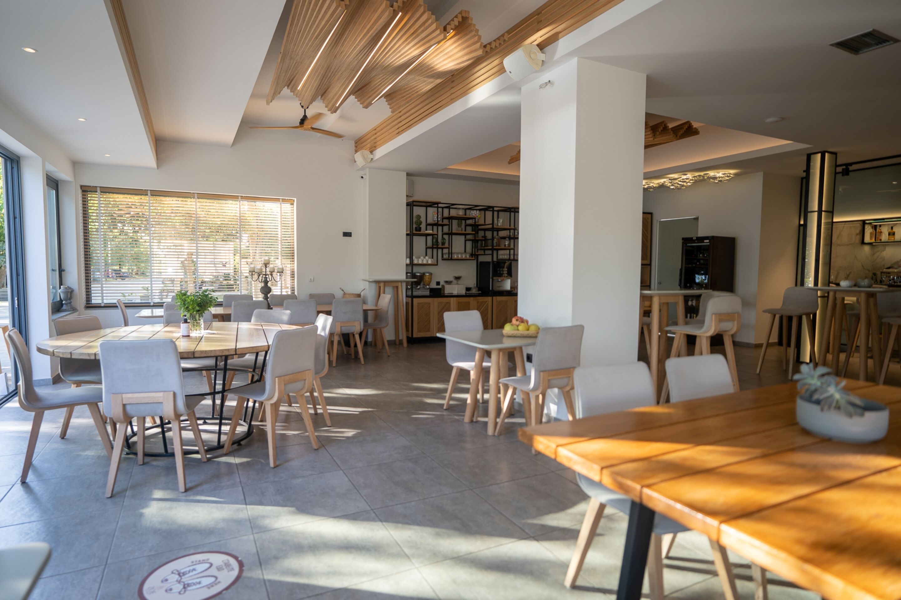 A spacious dining area with multiple tables and chairs, a kitchen in the background, and large windows allowing natural light.