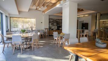 A spacious dining area with multiple tables and chairs, a kitchen in the background, and large windows allowing natural light.