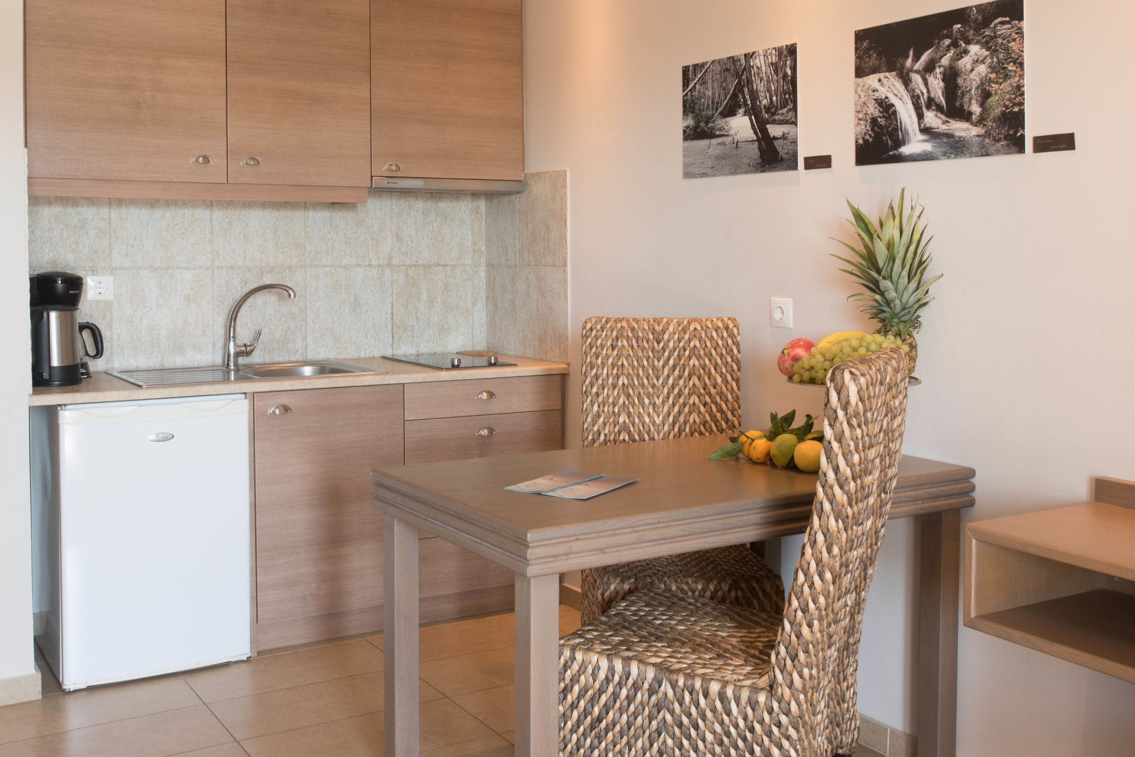 Cocina compacta con un lavamanos, cafetera y un refrigerador pequeño. Hay una mesa de comedor con una silla tejida y una cesta de frutas encima.