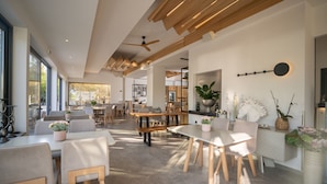 Un vaste espace dédié à la salle à manger, avec des tables et des chaises en bois, un ventilateur de plafond et de grandes fenêtres qui laissent entrer la lumière naturelle.
