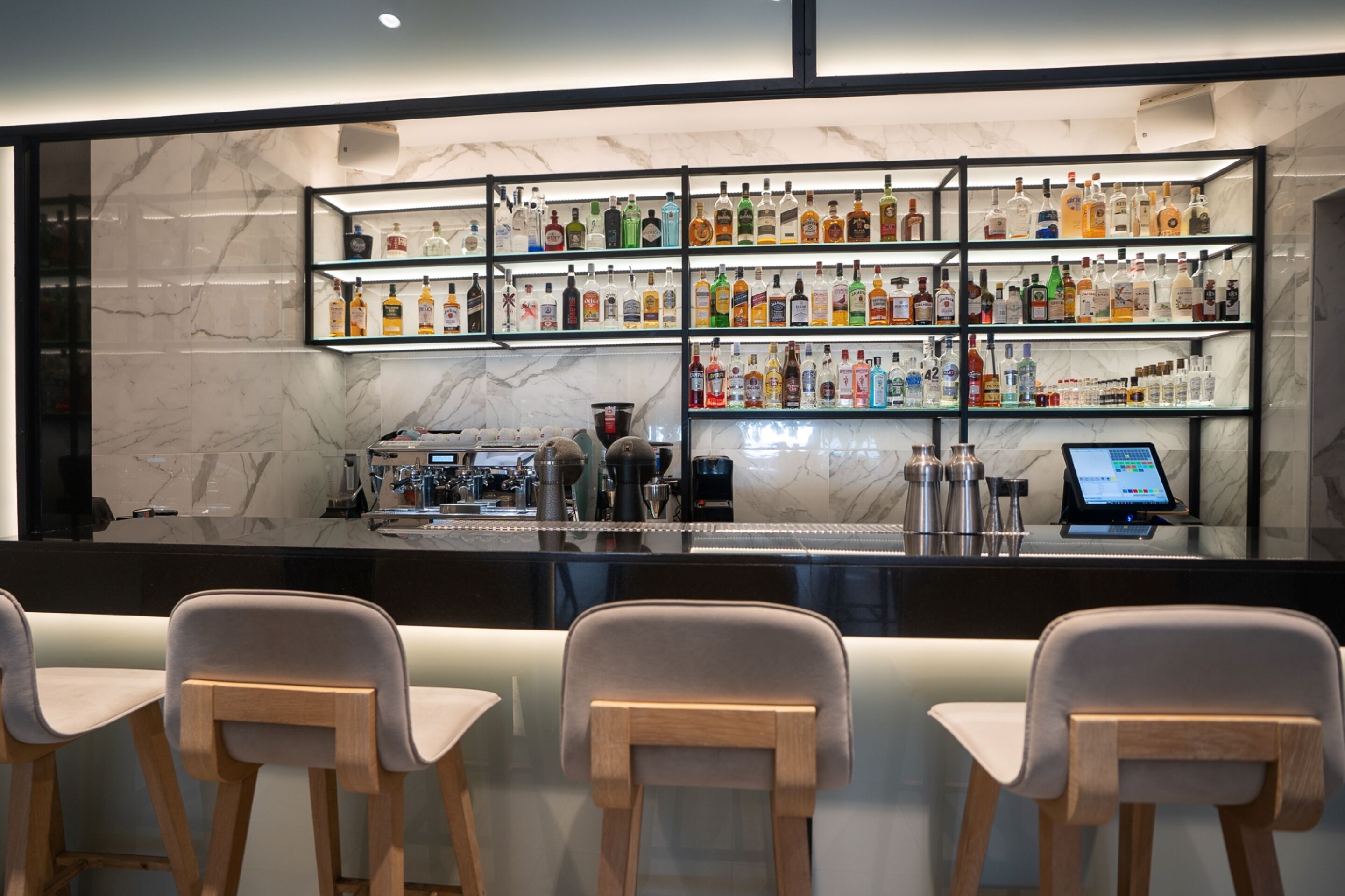 Un espace bar avec plusieurs étagères remplies de bouteilles d’alcool, un appareil à café et un terminal de paiement électronique. Trois chaises en bois sont placées devant le bar.