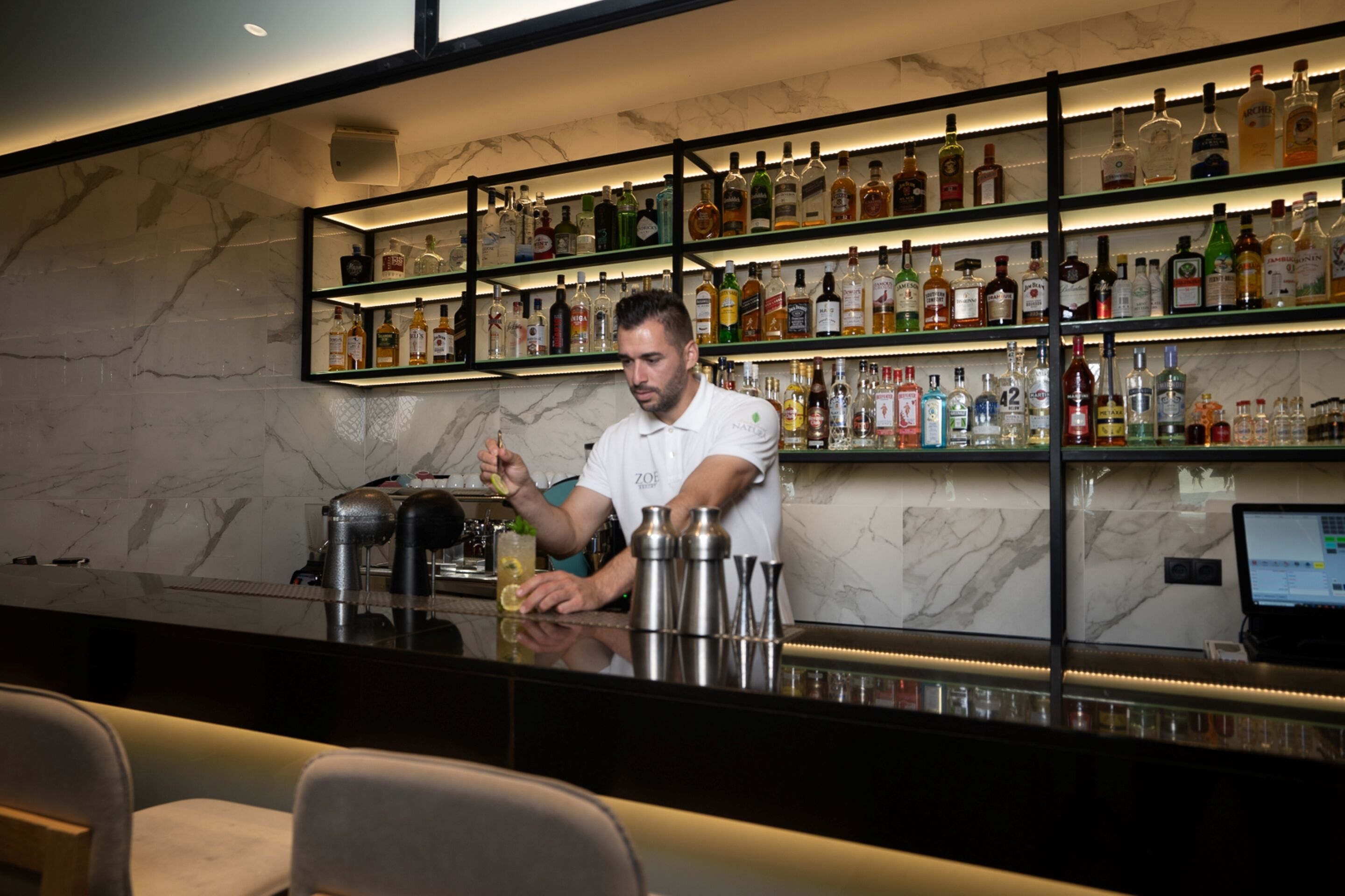 A bar area with a bartender preparing drinks, shelves of liquor bottles, and a laptop on the counter.