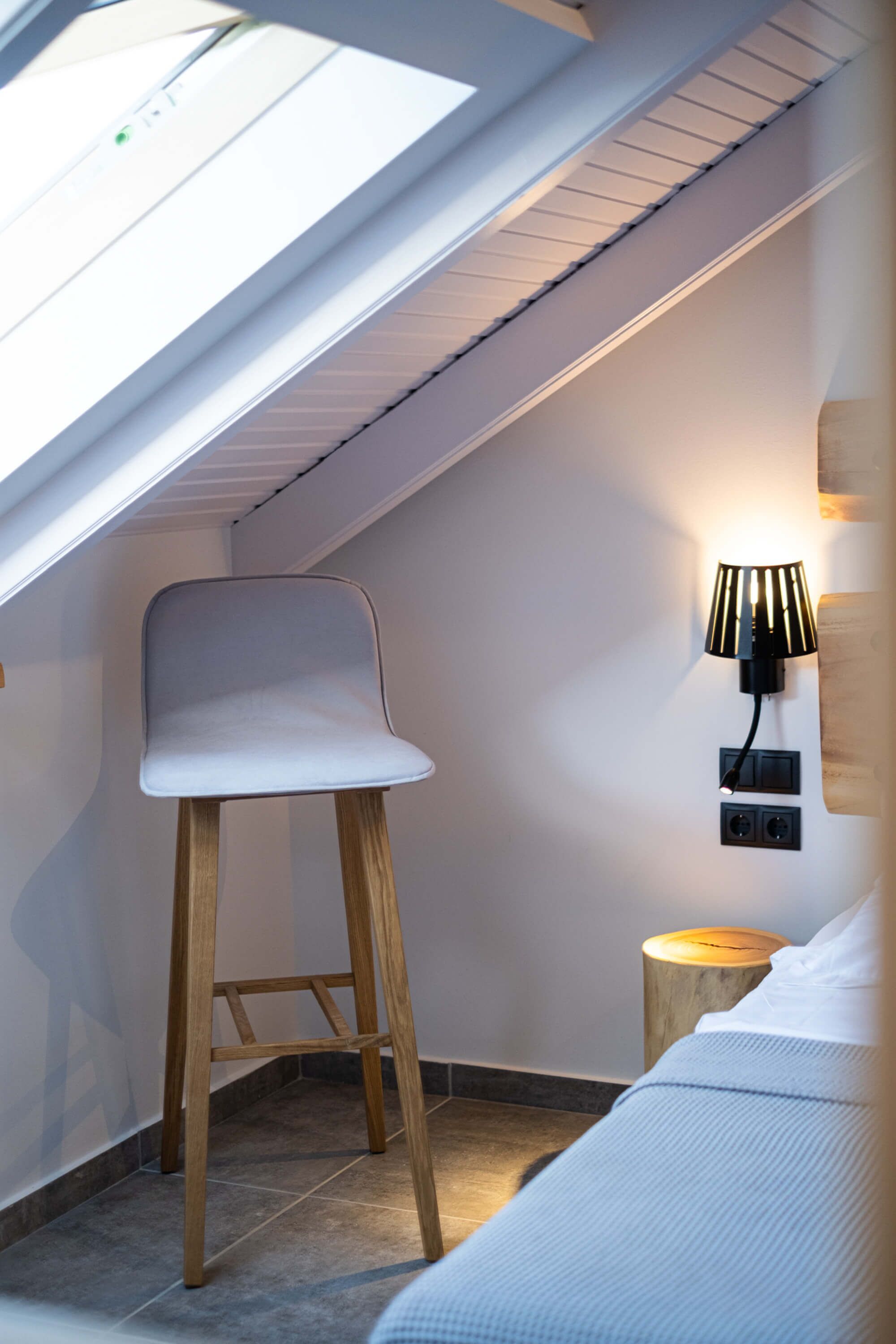 Ein kleiner, moderner Raum mit Oberlicht, einem Holzschemel, einem Nachttisch mit Lampe und einem Bett.