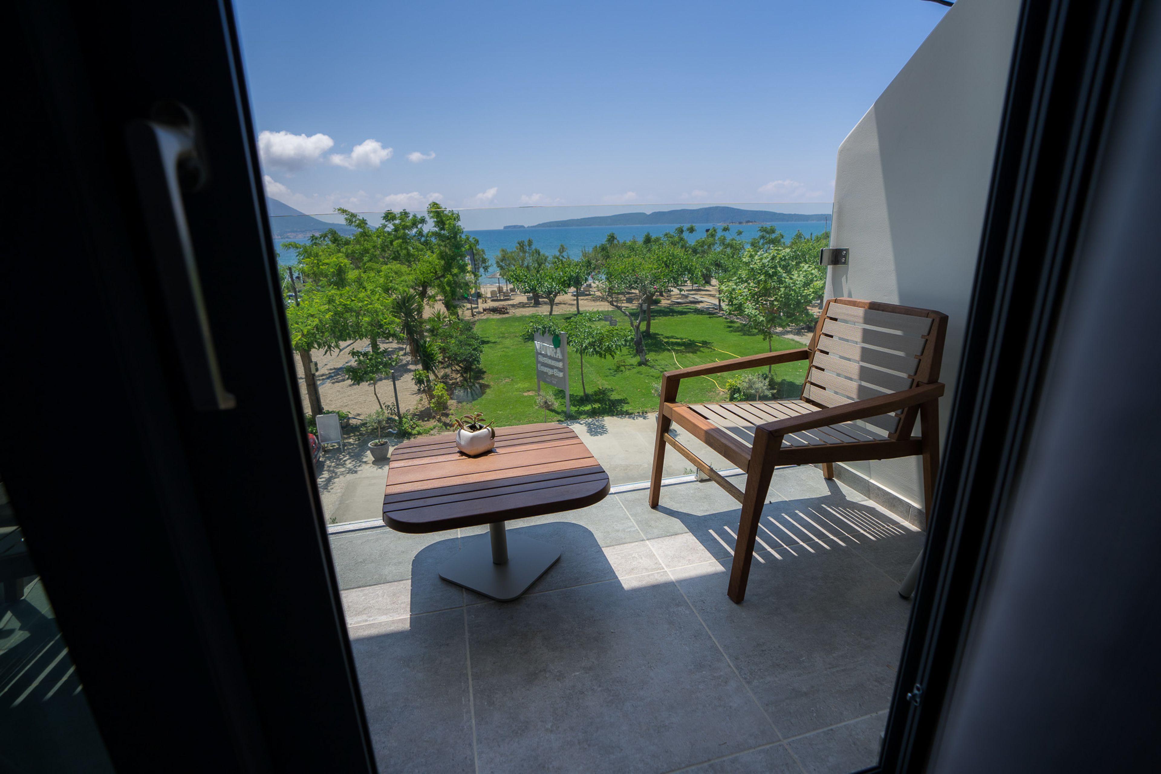 A balcony with a wooden chair and table, overlooking a landscaped garden and a distant view of the sea and mountains.