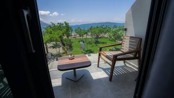 A balcony with a wooden chair and table, overlooking a landscaped garden and a distant view of the sea and mountains.