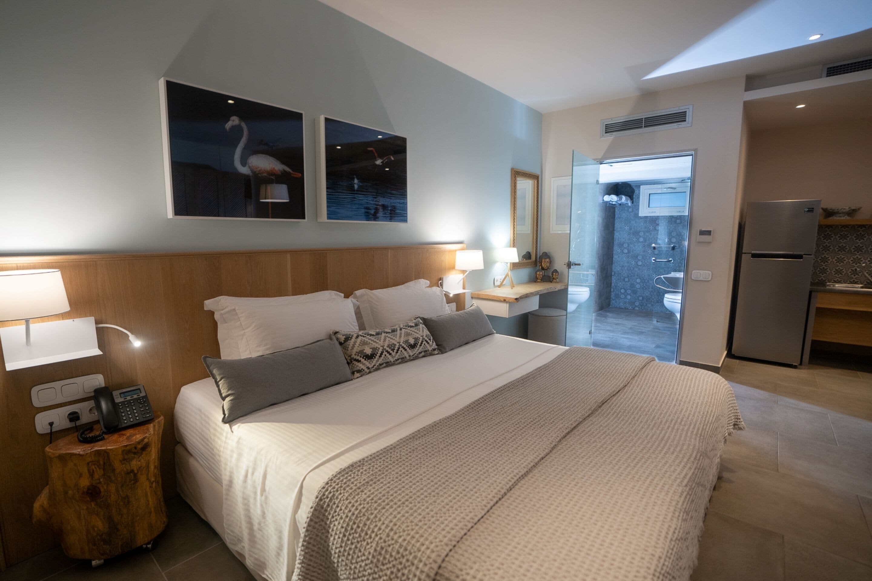A modern hotel room with a large bed, bedside lamps, a wooden nightstand, a mirror, and a view of the bathroom and kitchen.