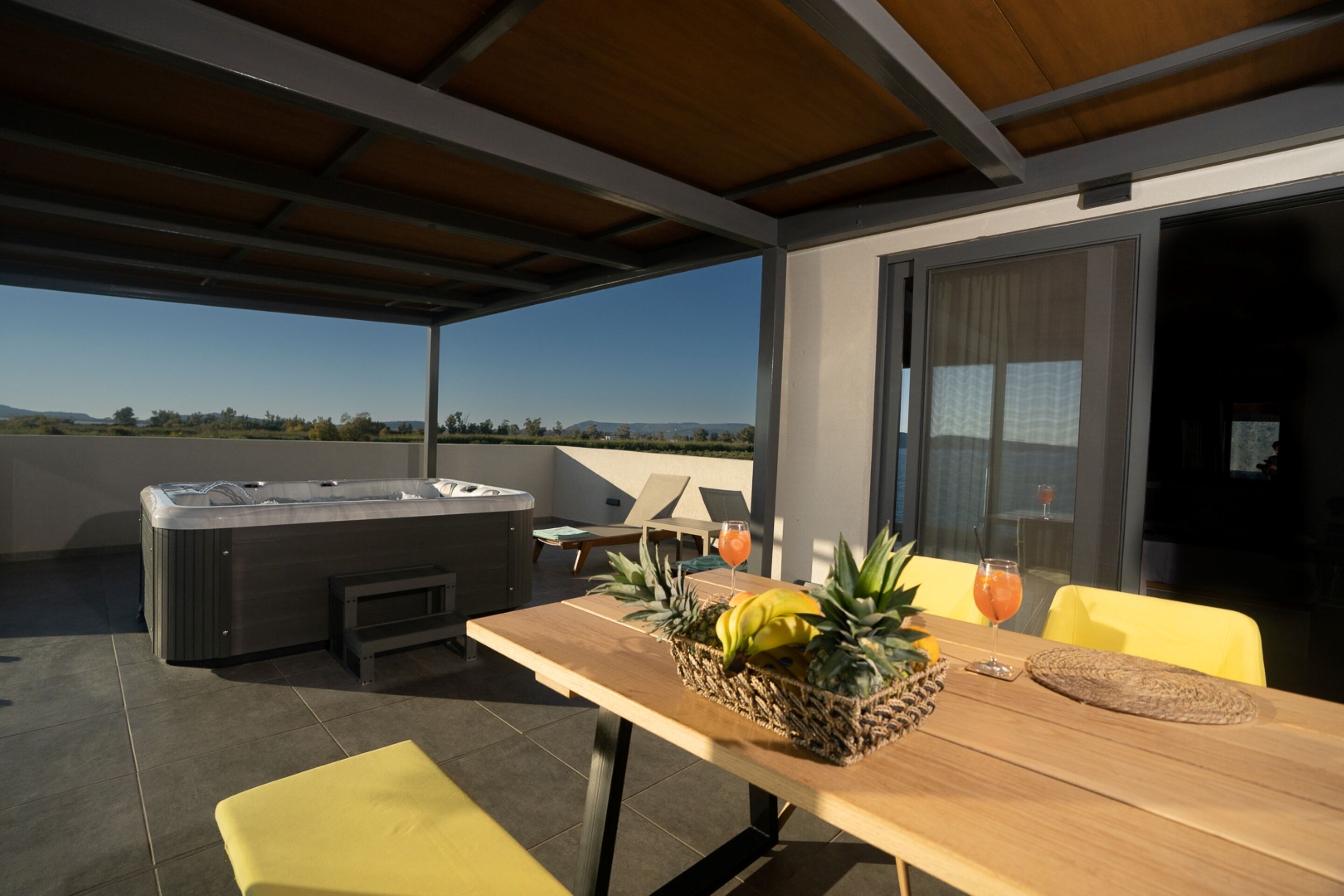 A covered outdoor area with a hot tub, a wooden table with a fruit basket, and yellow chairs.