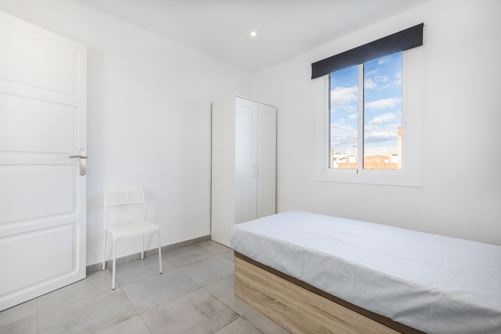 A bedroom with a bed, a chair, a wardrobe, and a window with a city view.