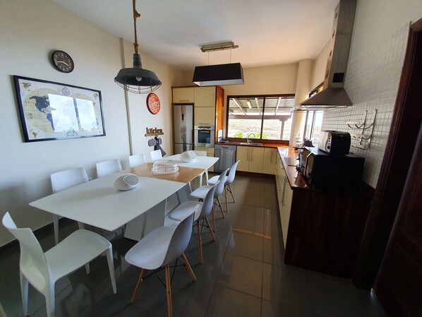 A modern kitchen with white cabinets, a dining area with a white table and chairs, a large window with a view, and a clock on the wall. - Gran Villa Vilena (Tijoco Bajo Adeje)
