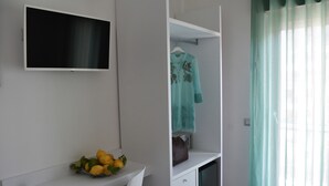 A modern room with a flat-screen TV mounted on the wall, a white desk with a bowl of lemons, a hanging garment, and a window with sheer curtains. - Vera Gioia Rooms de Charme (Sorrento)
