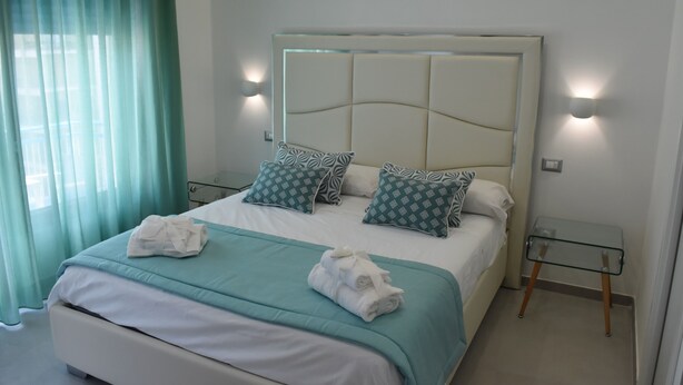 A neatly made bed with a teal bedspread, two patterned pillows, and two rolled towels. There is a glass side table with a lamp beside the bed. - Vera Gioia Rooms de Charme (Sorrento)