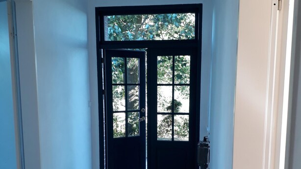 Un pasillo con una puerta negra que da a una habitación luminosa con vistas a árboles.