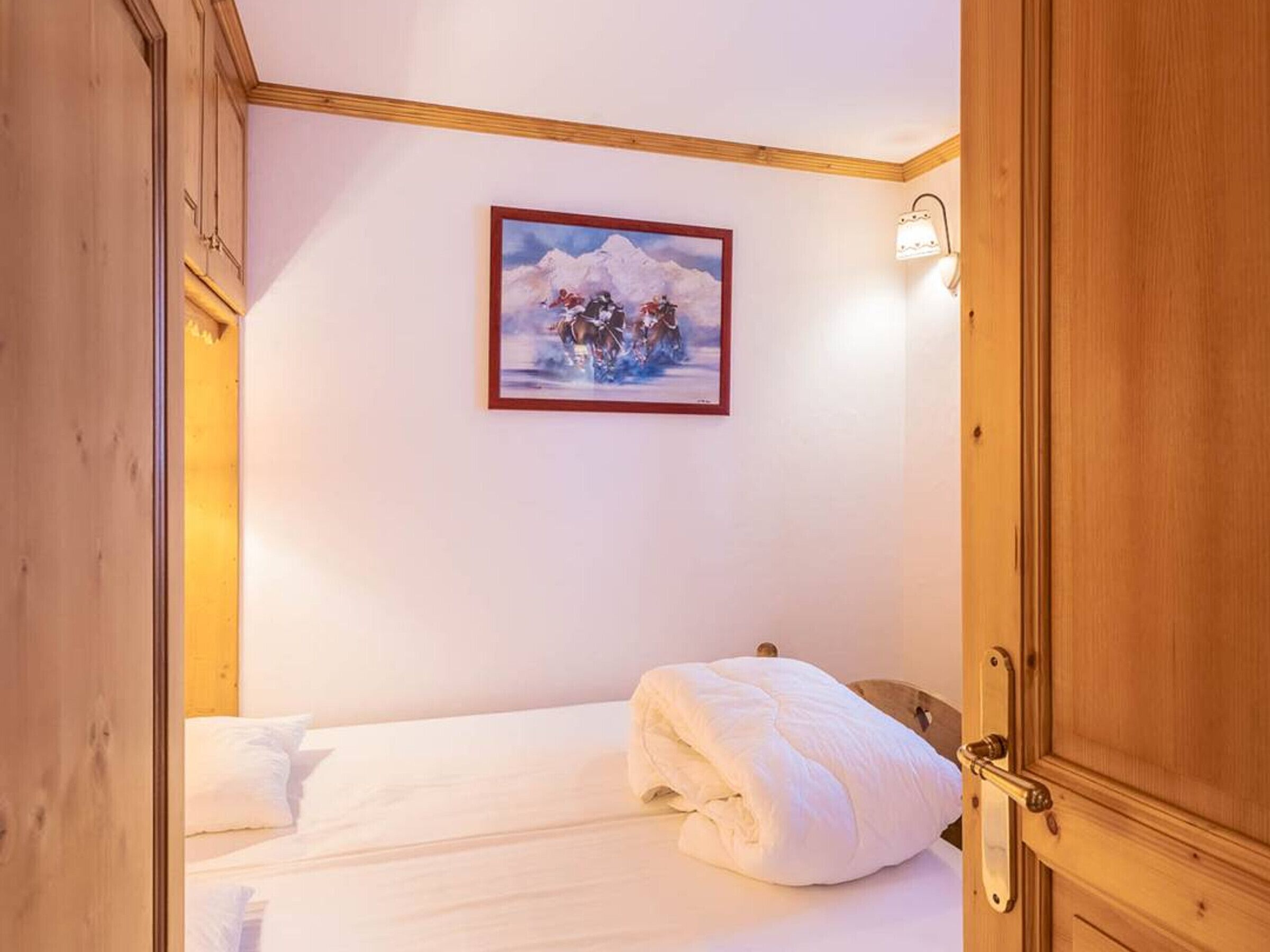 A hotel room with a bed, a painting of a snowy mountain scene, and a wooden door.