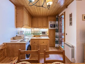 A compact kitchen with wooden cabinets, a washing machine, a dining table with chairs, and a window with a curtain.