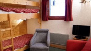 A bunk bed with a red patterned bedspread, a grey armchair, a red sofa, a television on a wooden stand, and a window with red curtains.
